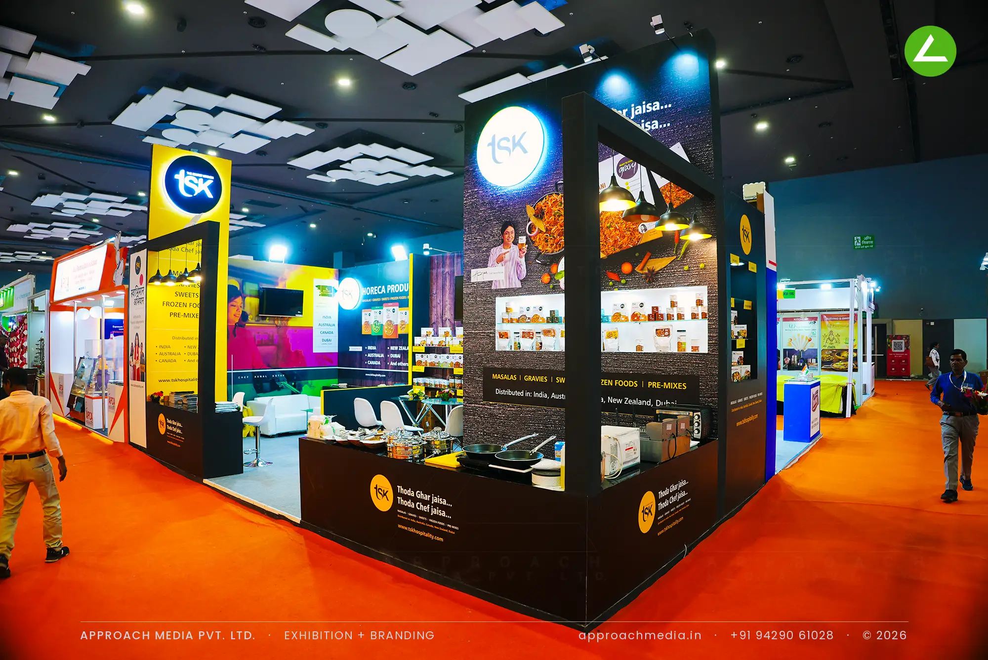 TSK exhibition stall design at Aahar New Delhi with dark plank walls, backlit round logo disc and a live cooking counter