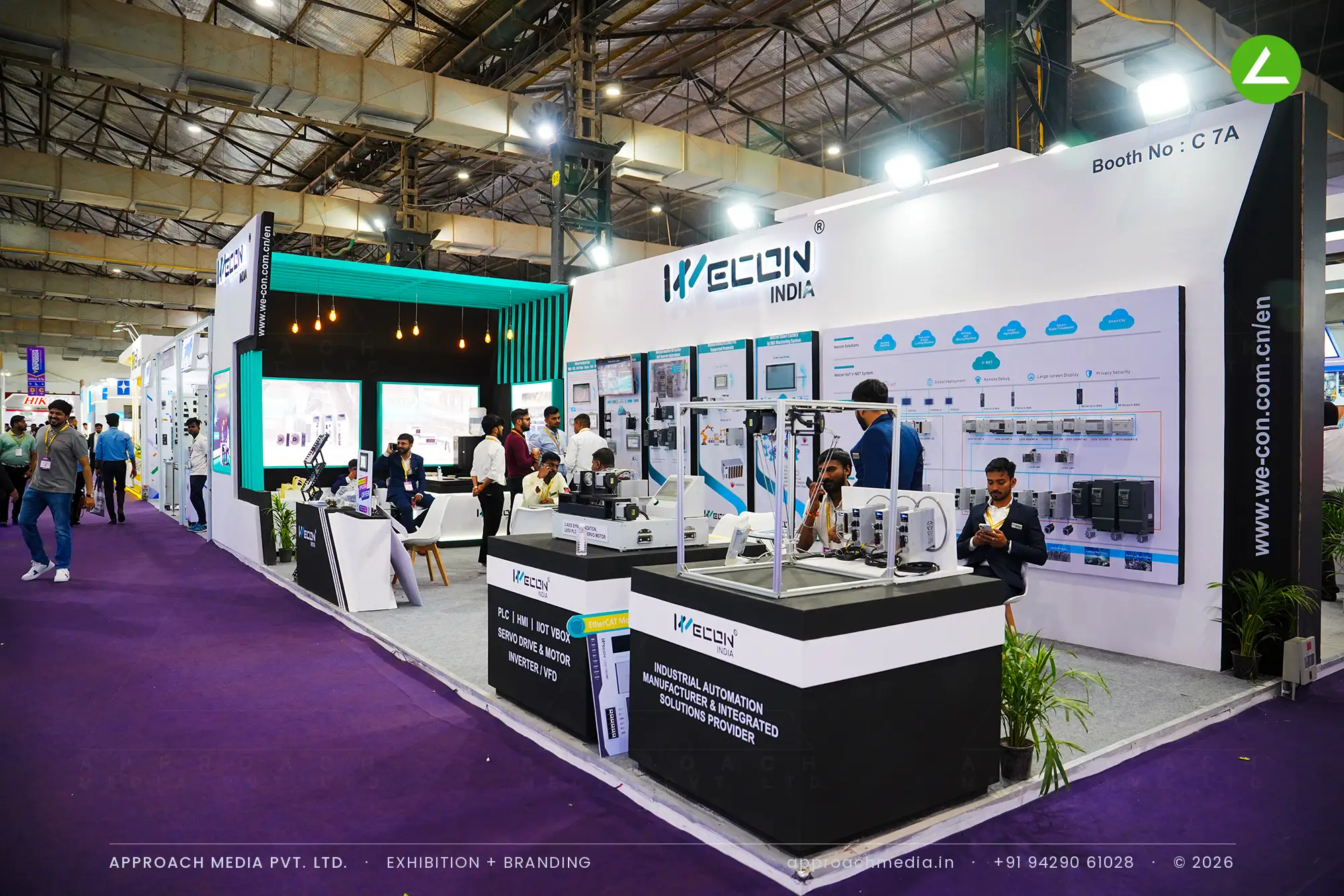 White demo counters with black tops stand in front of Wecon India's printed controller wall, below a teal slatted canopy.