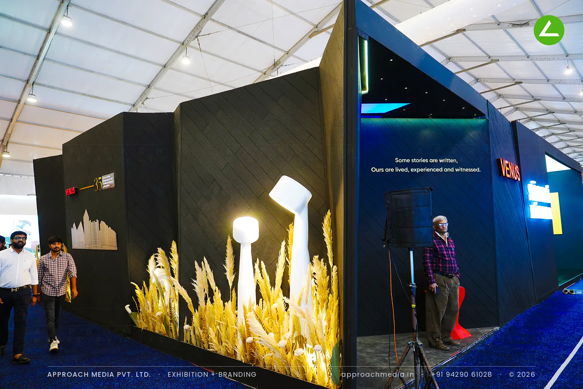Venus exhibition stall design at GIHED Property Show Ahmedabad: dark faceted walls, red neon sign and uplit dried grass